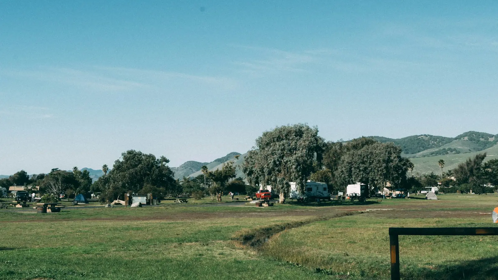 Grasveld van een camping met caravans en tenten tussen de bomen, met heuvels op de achtergrond